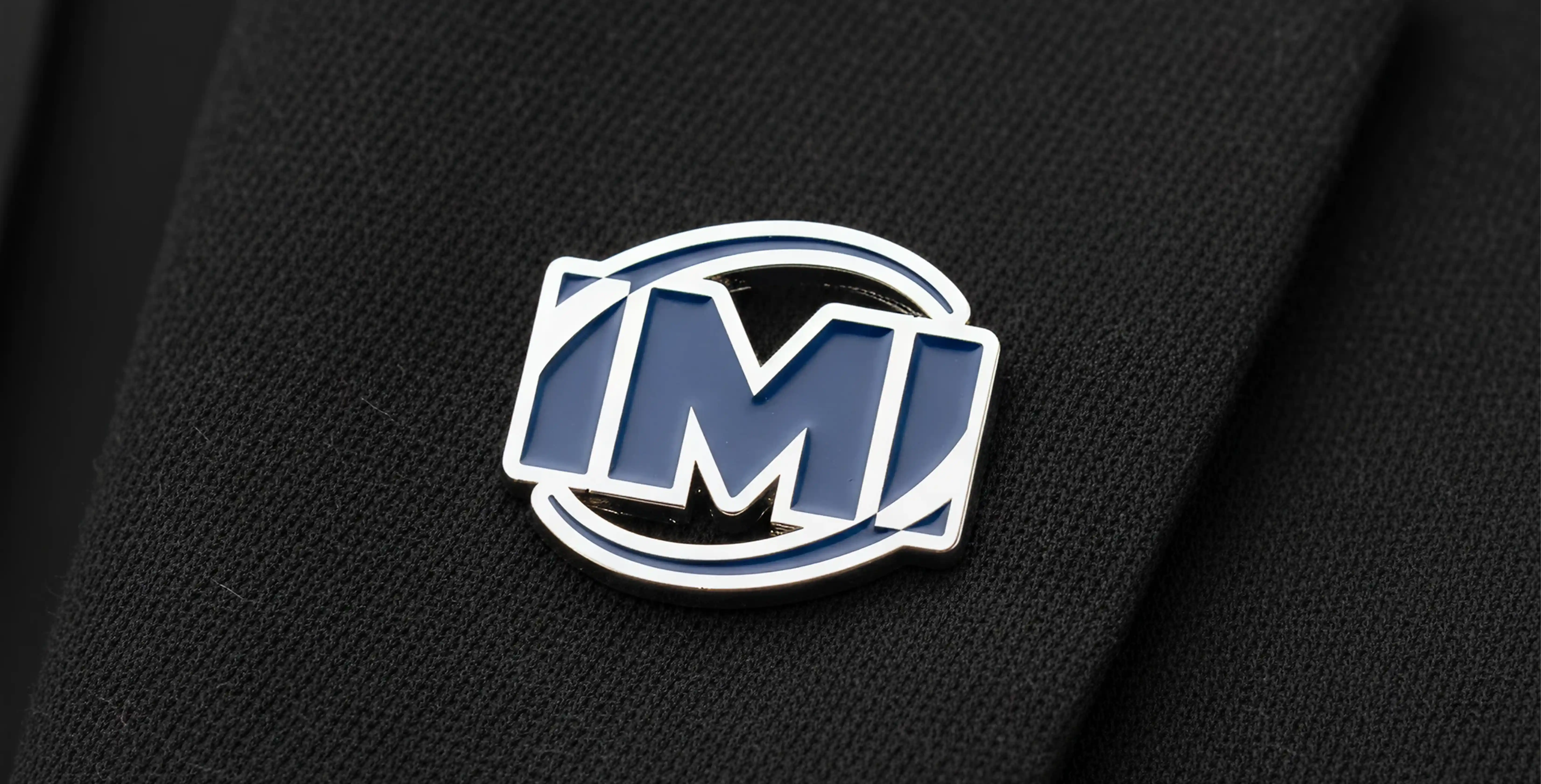 Bl&aring; och silver emalj "M"-logotyp n&aring;lm&auml;rke p&aring; svart tyg.